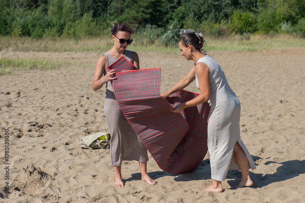 Fototapeta premium Two Caucasian women spread a blanket on the beach for a picnic. 