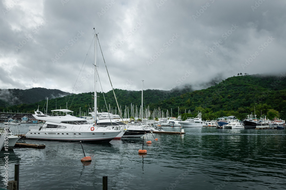 Fototapeta premium Calm marina filled with boats and yachts, surrounded by misty green hills and overcast sky creating serene atmosphere in Trinidad and Tobago