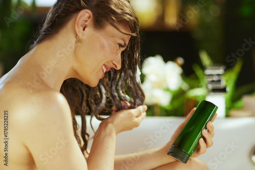 A smiling young adult woman with long, wet hair, applies hair product while holding a dark green bottle. The serene setting with blurred green foliage & white flowers conveys contentment & self-care.