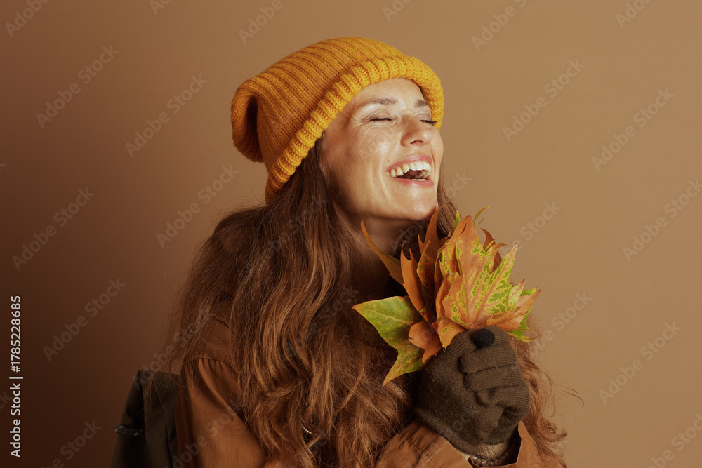 Fototapeta premium A woman, wearing a yellow beanie and brown jacket, laughs with her eyes closed while holding a fan of vibrant autumn leaves, perfectly capturing a joyful and festive fall mood.