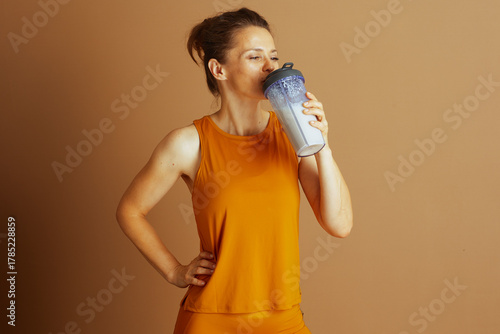 A focused woman in mustard yellow athletic top drinks from protein shaker in profile, hand on hip. Against light brown background, she embodies health, active living and post-workout hydration