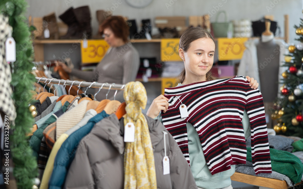 Fototapeta premium In sales area of store during Christmas sale, teen girl customer chooses coat or jacket. Creating capsule wardrobe, branded clothing at affordable prices. Modern urban fashion, casual practical