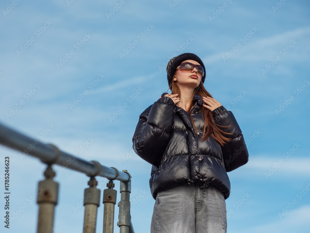 Obraz premium Young teenager girl in black jacket and grey jeans is standing on top of a stair with metal barrier, blue cloudy background. Model is wearing hat and sunglasses and enjoys warm sunny winter day.