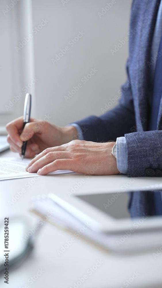 Fototapeta premium Closeup of businessman hands signing a contract with elegant pen, wearing a blue suit and a wristwatch, sitting at a white desk in office. Business people concept