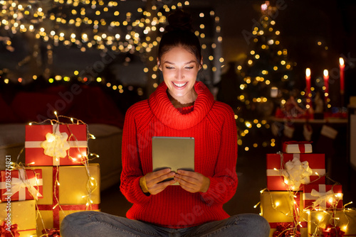 Satisfied woman using digital tablet, sitting in decorated room with Xmas gifts and garlands, spending Christmas time on internet ordering presents