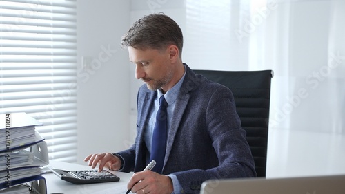 Professional businessman calculating finances, signing paperwork, wearing tailored suit while working at modern office desk. Audit and taxes in business