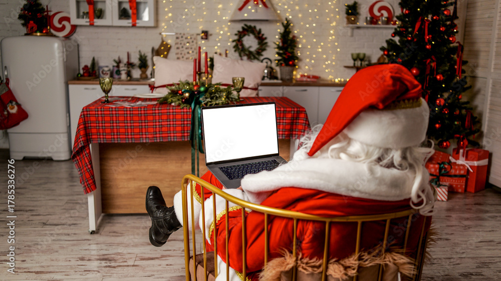 Obraz premium Santa Claus relaxing with a laptop in a festive Christmas decorated kitchen