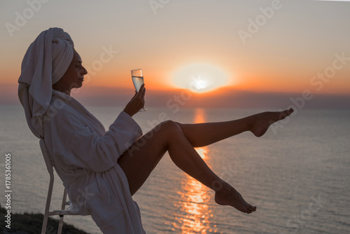 A woman in a white robe and a towel on her head drinks water, champagne from a glass against the sunset at the hotel. Sea, beach, vacation, sunscreen, warm weather, travel. High quality photo