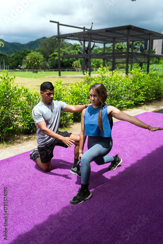Latin american personal trainer assisting a woman with her lunge workout, lifting a kettlebell on a purple exercise mat outdoors