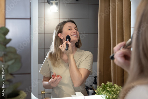 A woman applies makeup in the bathroom, using a powder brush in front of a lighted mirror. Morning beauty rituals, self-care, and creating a flawless look