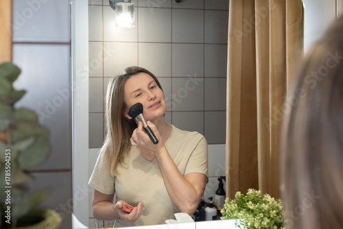 A woman applies makeup in the bathroom, using a powder brush in front of a lighted mirror. Morning beauty rituals, self-care, and creating a flawless look