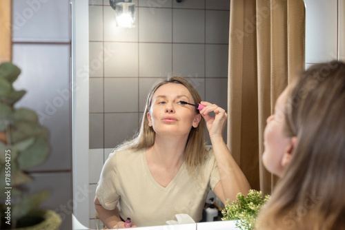 A woman applies makeup in the bathroom, using a powder brush in front of a lighted mirror. Morning beauty rituals, self-care, and creating a flawless look