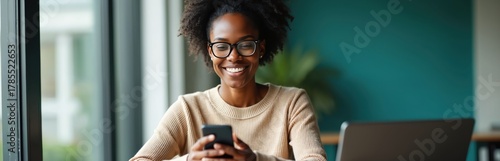 Wallpaper Mural Smiling woman wearing glasses uses phone in office setting. She looks happy while checking messages on mobile device, laptop nearby. Business professional enjoys break time in modern workspace. Torontodigital.ca