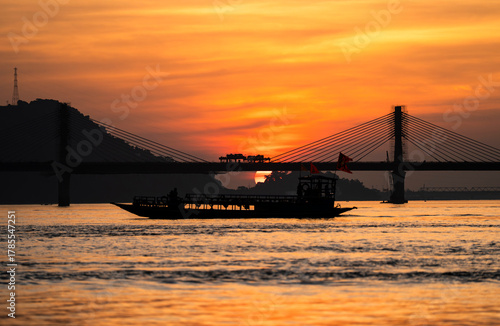 A boat sails on the river Brahmaputra during  sunset, with the silhouette of a cable-stayed bridge and hills in the background