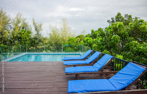Tropical resort rooftop swimming pool deck with wooden lounge chairs. Row of empty blue sunbeds on a modern rooftop deck overlooking an exclusive infinity pool and lush green tropical surroundings.