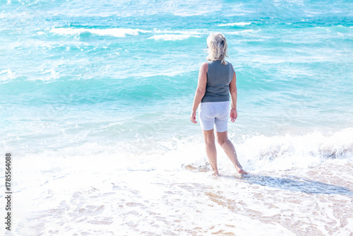 An elderly woman taking a walk by the sea. A happy lady in sportswear stands in the beautiful turquoise water and looks into the distance. Activity, peace, and freedom. Back view.