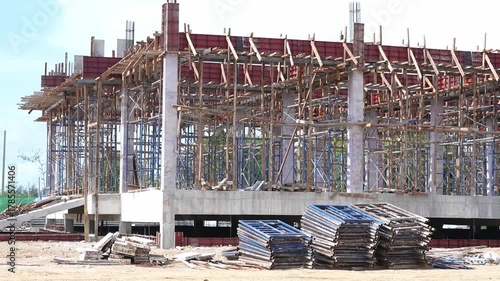 Construction site with concrete structure, scaffolding, stacked metal formwork panels, steel beams, and wooden planks, showing progress of building development and framework bright daylight