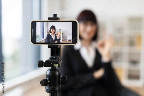 Caucasian woman wearing formal attire recording video blog using smartphone on tripod in modern office setting. Concept themes include business networking, educational content creation.