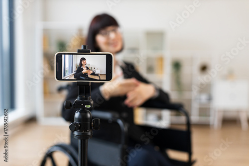 Caucasian woman seated in wheelchair recording blog on smartphone about business topics. Bright room with tripod and background bookshelf provide professional and creative environment for filming.