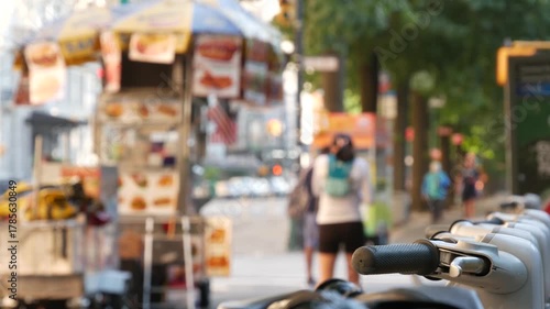 Manhattan Midtown street New York City, United States. Rental bicycle parking, Central Park West summer trees greenery. Bike for rent in row. People pedestrian on sidewalk pavement. Hot dog fast food.