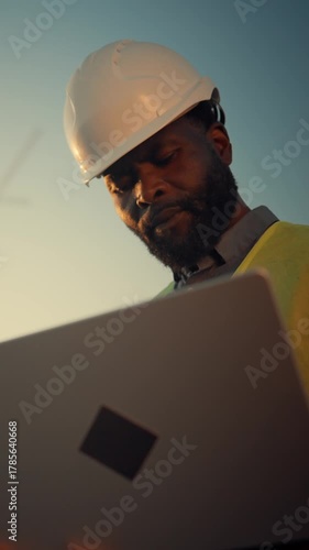 Architect or inspector with laptop in construction site, portrait of builder professional. Vertical video with serious black man controlling process of building new district in modern city, innovation