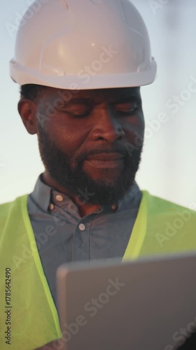 Bearded foreman viewing drawings on display of modern laptop in construction site. Vertical portrait view, modern innovation in building and development of cities, build new district in megapolis
