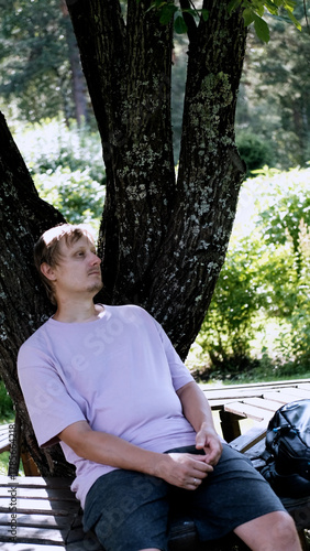 Relaxing man in a park on a bench in summer, enjoying the fresh air, vacation, or relaxing alone in the garden. A thoughtful, smiling, or calm man in nature, resting or waiting in a natural setting.