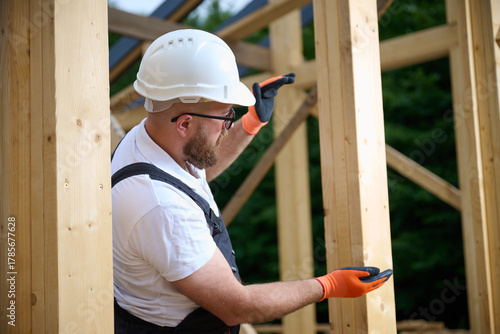 Construction worker checking wooden beam alignment at timber frame house site. Builder in safety gear holding helmet and using tools while working on wooden structure outdoors.
