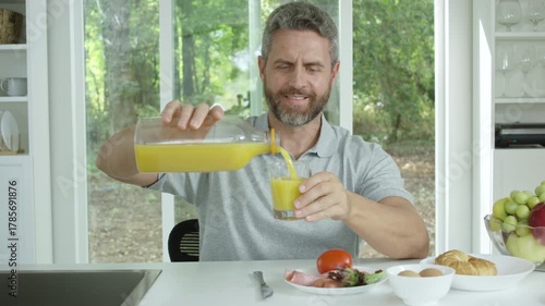 Man pouring orange juice at morning kitchen. Mature man pours fresh orange juice for breakfast. Middle aged nan eating breakfast in cozy kitchen.