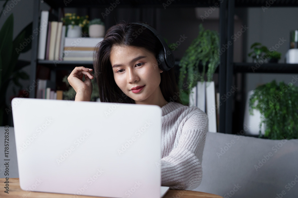 Naklejka premium Asian woman sits on sofa wearing headphone watching movie on laptop in living room