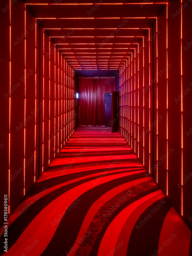 Fototapeta premium A vibrant, futuristic tunnel illuminated by an immersive grid of red LED lights, creating a captivating perspective. The floor features dynamic, flowing red and black stripes, leading towards a