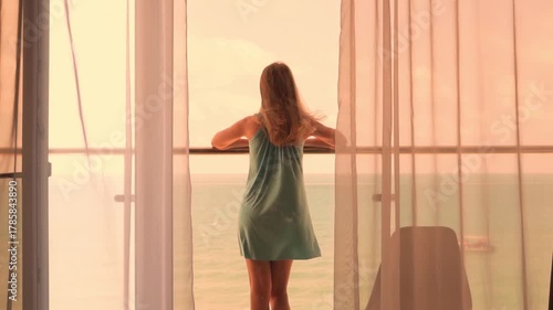 Woman Tourist Stands on the Terrace of a Apartment and Admires the Beautiful Sea View and Orange Yellow Sunset Sky. Dress Hair and Sheer Curtain Swaying in the Wind Slow Motion. Beachfront Real Estate
