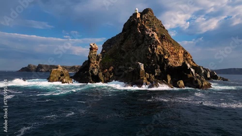 Aerial view of waves crashing against rugged rocks at Cape Point crowned by a white lighthouse under a blue sky, Cape Town, Western Cape, South Africa.