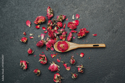 Small dried rose buds and petals scattered on a dark surface with a wooden spoon holding some buds.