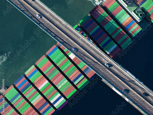 Aerial view of a massive cargo ship, its colorful containers contrasting with the dark blue sea, bisected by a concrete bridge bustling with tiny cars, Los Angeles, California, United States.