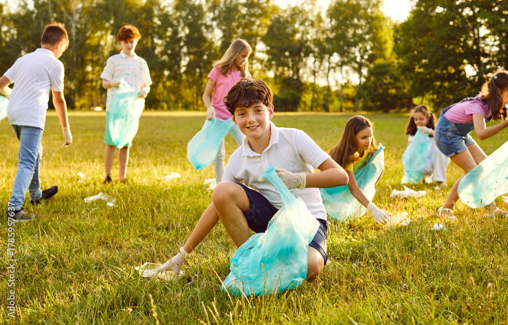 Fototapeta premium Portrait of a happy teenage boy with gloves and trash bag collecting plastic garbage in the summer park with a children volunteers on the background. Environmental pollution and ecology concept.