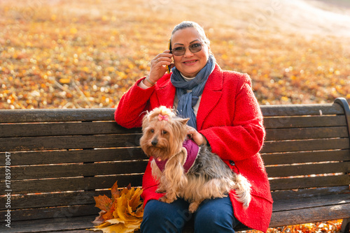 Fototapeta Woman sits on a bench in a park with her dog during autumn afternoon