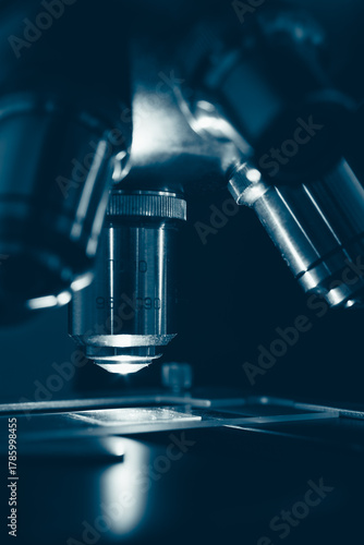 Macro photo of optical microscope lens focusing on sample slide under sterile laboratory lighting. Symbolizes technology, research, and medical discovery.