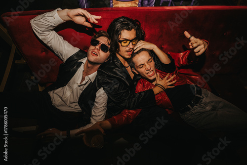 Schilderij op canvas A lively nightlife scene with three friends in sunglasses and leather jackets, posing energetically on a red couch