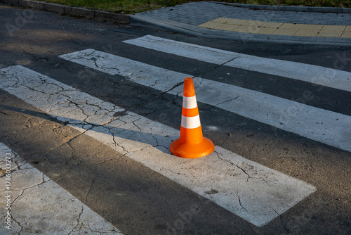 An orange traffic cone rests on a cracked crosswalk in an urban area, with sunlight casting shadows on the pavement