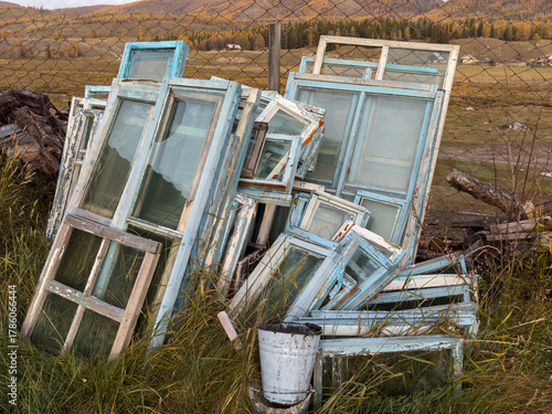 Painted window frames stacked beside fence in rural field with autumn hills. Weathered blue frames create rustic collage of glass and weathered wood.