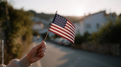 Fototapeta Naklejka Na Ścianę i Meble -  Hand holding a small USA flag while standing on a quiet morning street with soft sunrise light illuminating the surroundings — concept of patriotism, national pride, morning serenity, and lifestyle