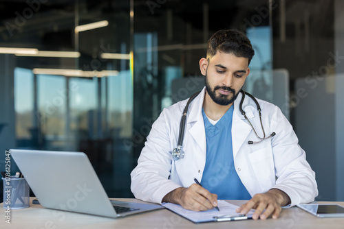 Young male physician wearing a lab coat and stethoscope working on healthcare documentation at a desk with a laptop, and tablet in a contemporary office setting