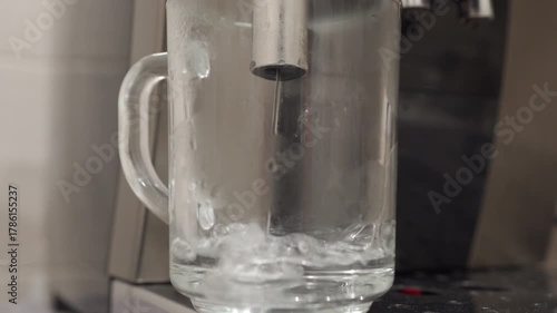 A metal coffee machine dispenser is pouring hot water into a glass mug. The liquid rapidly fills the cup, creating a continuous gushing and bubbling sound.