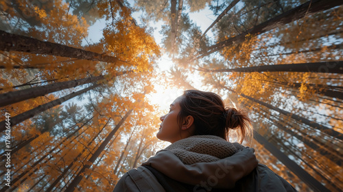 Woman in autumn clothing standing in a vibrant orange and yellow forest, looking up towards the bright sun shining through tall towering treetops.