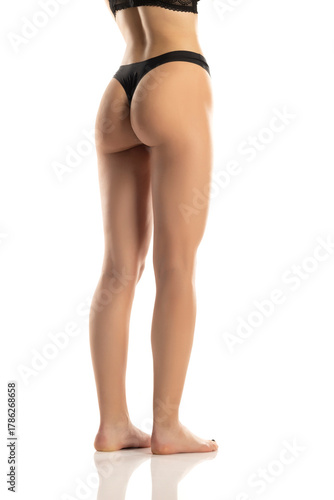 Rear view of a woman’s legs, buttocks, and feet wearing black lingerie standing barefoot with smooth skin, studio shot on a white background