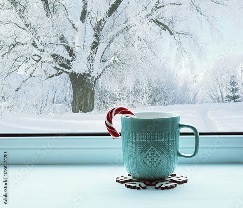 Winter background, green mug with candy cane on windowsill and winter forest landscape outside. Cozy winter still life with concept of spending winter time indoors