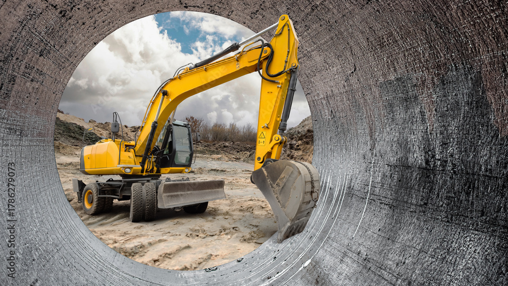 Fototapeta premium Heavy machinery is actively digging on a construction site, viewed through a large pipe that frames the scene against a cloudy sky. Loader, excavator
