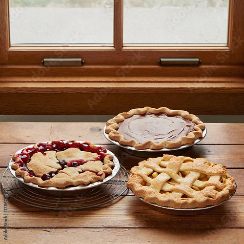 Three freshly baked homemade pies sit on a rustic wooden table, cooling by the window.