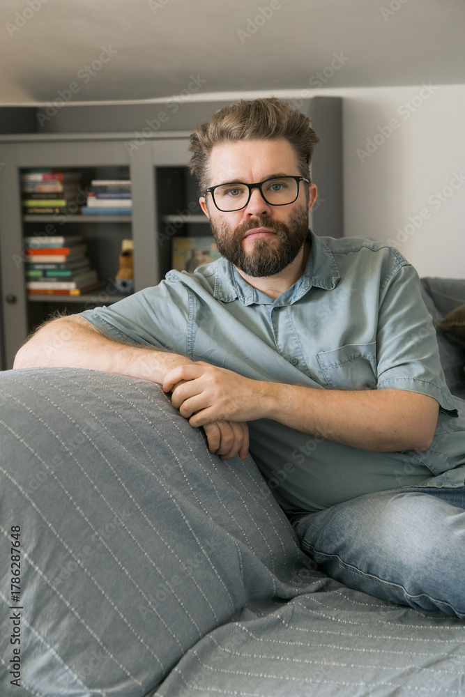 Fototapeta premium Man sitting on couch in home living room. Representation of relaxation, thoughtfulness, and calm everyday moment in domestic environment.